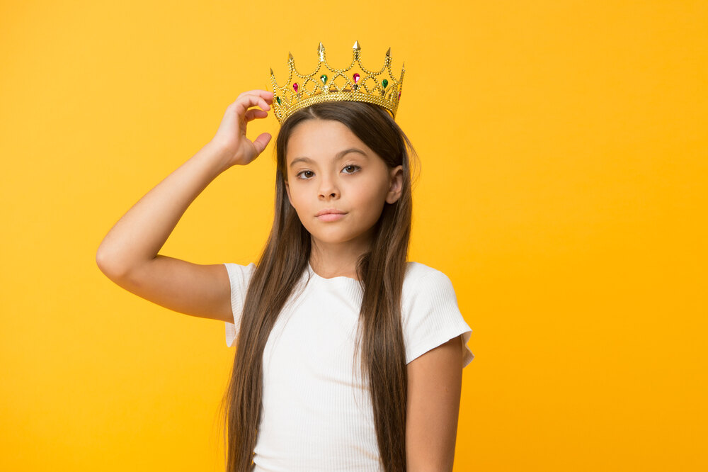 A young girl looks confidently into a camera, adjusting a pageant crown on her head.;